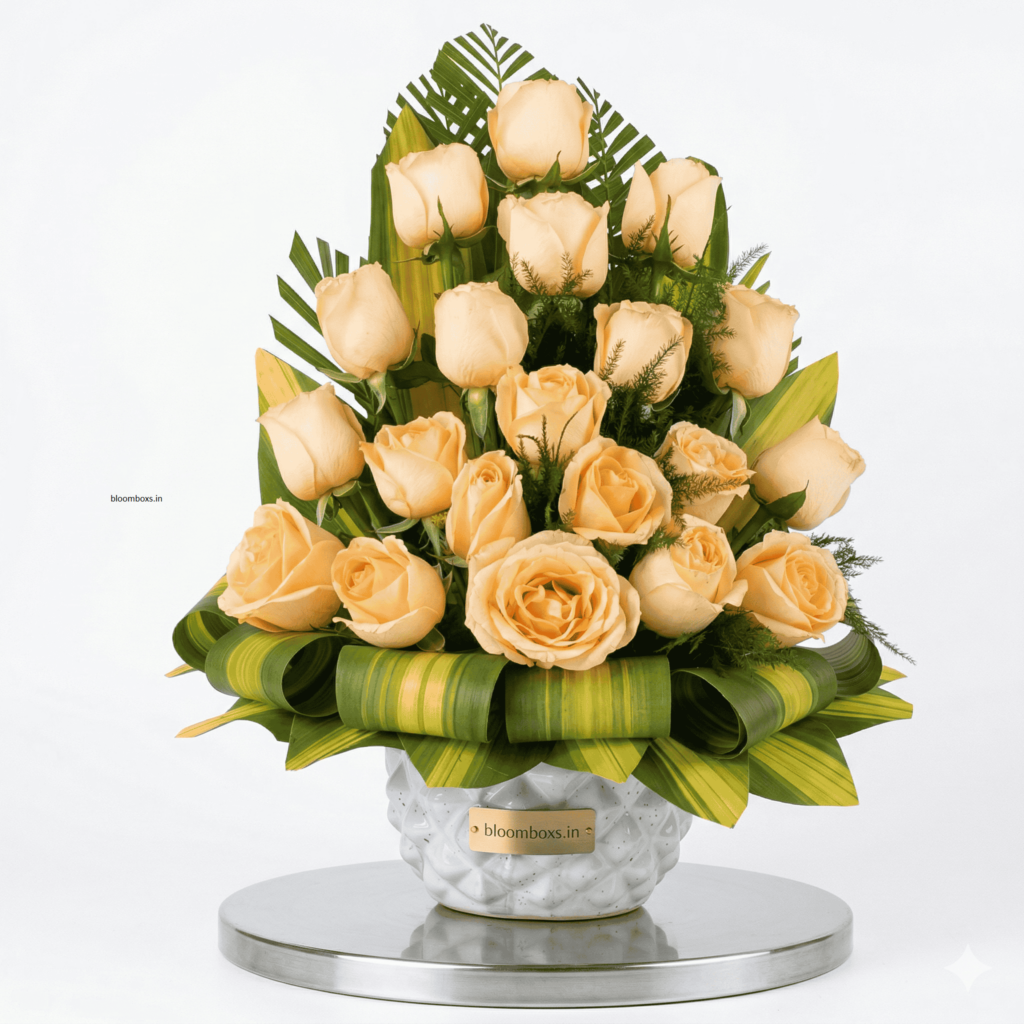"A luxury floral arrangement featuring two dozen peach-colored roses arranged in a dome shape, accented with tropical green leaves and fern fronds. The bouquet sits in a white textured ceramic vase placed on a round silver mirror base, with a gold 'bloombox.in' tag on the front."